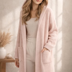 May include: A long, pale pink hooded cardigan with two front pockets. The cardigan is made of a soft, knitted material. The model is wearing a beige top and trousers. The cardigan is a stylish outerwear option.