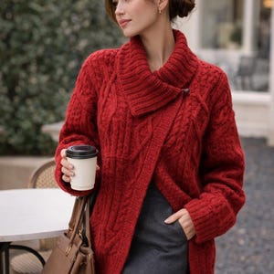 Puede incluir: Una mujer con un suéter rojo de punto trenzado con cuello chal y cierre de botón. Sostiene una taza de café blanca y un bolso de mano de cuero marrón. Lleva una falda gris.