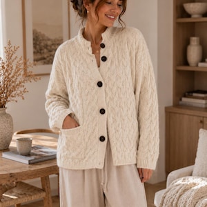 May include: A cream-colored cable knit cardigan featuring a mock neck, long sleeves, and a front button closure with five dark brown buttons. The garment includes two front patch pockets and is crafted with a textured vertical cable pattern.