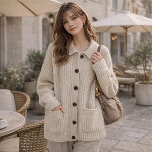Puede incluir: Un cárdigan beige con botones, cuello y dos bolsillos delanteros. El suéter tiene botones marrones oscuros y está hecho de un material de punto acanalado. Se ve un bolso beige a juego con una correa de cadena.