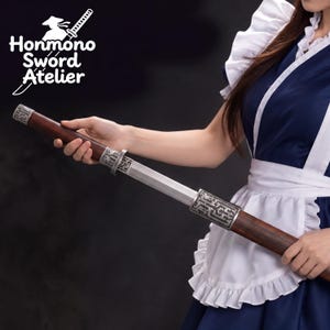 May include: A sword with a dark wood handle and scabbard featuring intricate silver-toned metal fittings with engraved patterns. The blade is partially unsheathed, revealing a metallic finish. The logo Honmono Sword Atelier is visible.