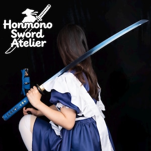 Puede incluir: Una katana azul y plateada con mango negro. La espada la sostiene una persona con un vestido azul y blanco. La imagen incluye el texto "Honmono Sword Atelier" en blanco.