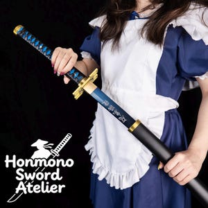 Pode incluir: Uma espada katana azul e preta com detalhes dourados. A pega apresenta um padrão de losangos azuis. Caracteres japoneses são visíveis na lâmina. A espada é segurada contra um fundo escuro. O logótipo Honmono Sword Atelier está no canto inferior esquerdo.