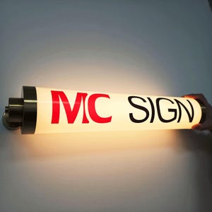 May include: A cylindrical illuminated sign with the text "MC SIGN" in red and black. The sign is mounted on a wall and has a warm, glowing light. The sign is held by a person's hand.
