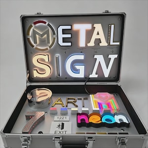May include: A silver metal sample case containing various illuminated signage elements. The lid displays the words METAL SIGN in large, backlit letters. The base features a variety of metal and neon letter samples, including the word ART, a number 7, and a neon sign.