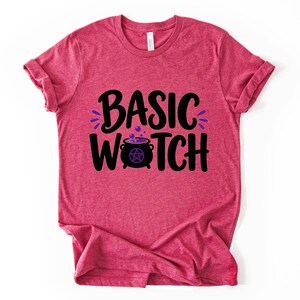 May include: A heathered red short-sleeve t-shirt featuring the black text BASIC WITCH printed on the front. The letter I in WITCH is replaced by a black cauldron graphic containing a purple pentacle symbol and purple bubbles rising above it.
