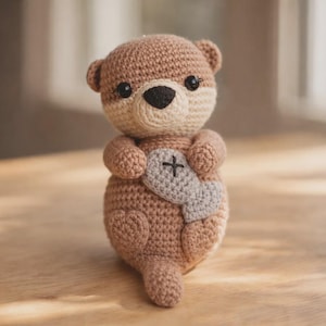 May include: A handmade crochet otter plushie in shades of brown and cream. The otter has a gray heart-shaped detail with a black cross stitch. The plushie has small black eyes and a black nose. The otter is sitting on a wooden surface.
