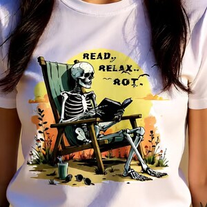 White t-shirt featuring a skeleton reading a book while relaxing in a beach chair. The graphic includes the text "READ, RELAX, ROT" with a sunset background, sunglasses, and a drink.