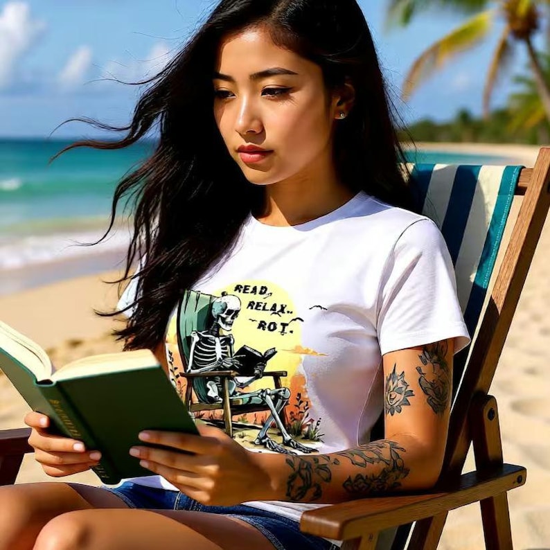 White t-shirt with a graphic of a skeleton reading on a beach chair, with the text "READ, RELAX...-ROT." The shirt is worn by a person sitting in a wooden beach chair, holding a green book. The background is a beach scene.