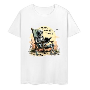 White t-shirt featuring a skeleton relaxing in a beach chair, reading a book. The graphic includes the words "READ, RELAX, ROT." Sunglasses and a drink are next to the chair. The background shows a sunset and birds.