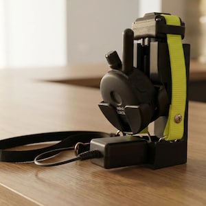 May include: A black dog training collar with a yellow strap is mounted on a black charging station. A black strap and charger are also visible. The setup is on a wooden surface.
