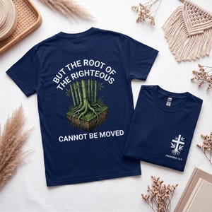 以下が含まれることがあります： 背面に土の中の竹の根のグラフィックと「BUT THE ROOT OF THE RIGHTEOUS CANNOT BE MOVED」の文字、前面の胸元に「PROVERBS 12:3」の文字が入った小さな白い十字架のグラフィックがプリントされたネイビーブルーのTシャツ。