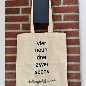 Könnte beinhalten: Beige Tragetasche mit schwarzem Text "vier neun drei zwei sechs" und "Wellingholzhausen" in Schreibschrift. Die Tasche hängt an einem dunkelblauen Halter vor einer roten Backsteinmauer.