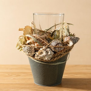 May include: A decorative arrangement with a clear glass vase inside a small, weathered grey metal bucket. The vase is filled with seashells, dried plants, and rope, creating a coastal theme. The bucket sits on a wooden surface.