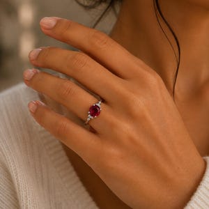 May include: A silver ring featuring a round, deep red gemstone at the center, flanked by two smaller, clear, round-cut stones on each side. The band is thin and metallic, showcasing a polished finish that complements the vibrant red center stone.