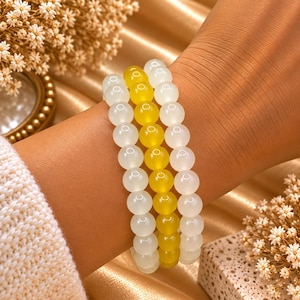 May include: A set of three beaded bracelets featuring smooth, round beads. Two bracelets consist of translucent white beads, while the center bracelet is composed of vibrant yellow beads. The beads are strung on an elastic cord for a flexible fit.