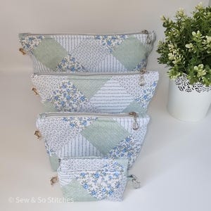 May include: Four quilted cosmetic bags in different sizes. The bags have a patchwork design with blue floral and green diamond patterns. Each bag has a silver zip and a small charm. The bags are stacked on top of each other.