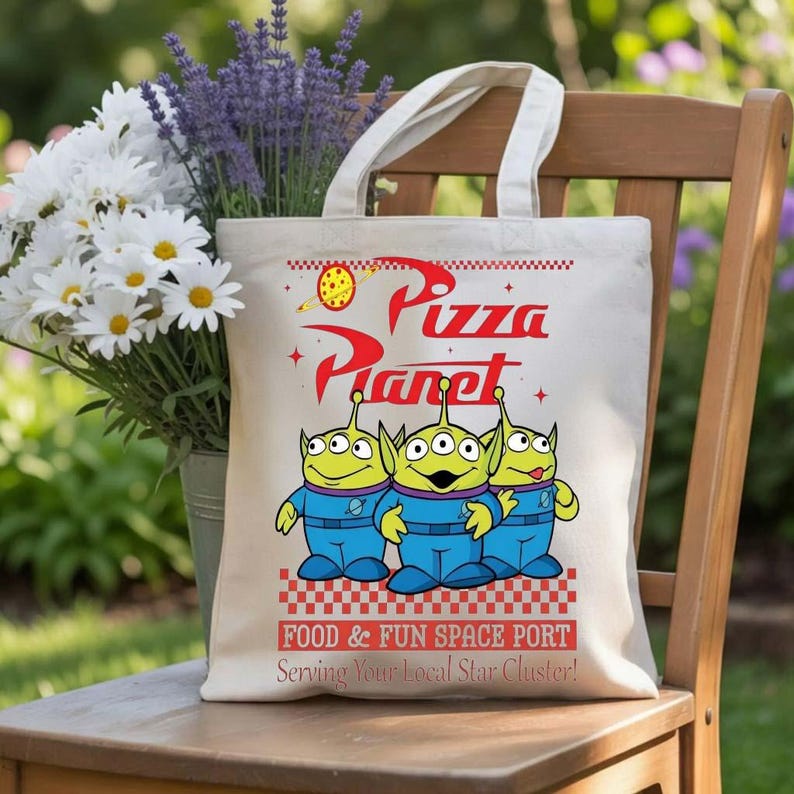 A white tote bag with a design featuring three alien characters from the Pixar movie "Toy Story" and the text "Pizza Planet" and "Food & Fun Space Port" printed on it. The bag is placed on a wooden chair, with a vase of white flowers in the background.