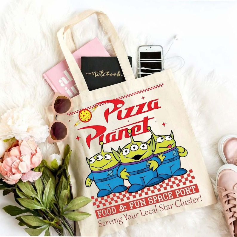 a white tote bag with a cartoon of three alien characters from the movie "Pizza Planet" and the text "Food & Fun Space Port" printed on it. The bag is placed on a white surface, surrounded by various items such as a smartphone, a notebook, sunglasses, and a pair of pink sneakers.