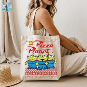 A woman is sitting on the floor with a tote bag featuring a design of three alien characters from the Pixar movie "Pizza Planet".