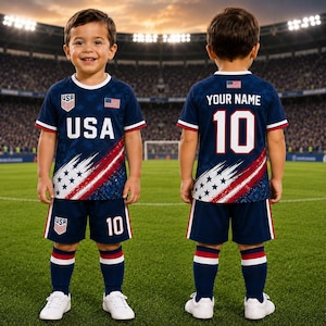 May include: A navy blue football kit featuring a white USA logo, the text USA, and a red, white, and blue star pattern. The back displays the text YOUR NAME and the number 10. The set includes a short-sleeved shirt, matching shorts, and football socks.