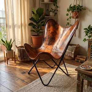 May include: A brown leather butterfly chair with a black metal frame. The chair has a distinctive shape and is placed in a room with plants and natural light. The leather has a distressed look.