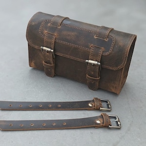 May include: A dark brown leather cylindrical bag featuring a distressed finish, a flap closure secured by two adjustable straps with metal buckles, and two separate matching leather straps with metal buckles positioned in front of the bag.