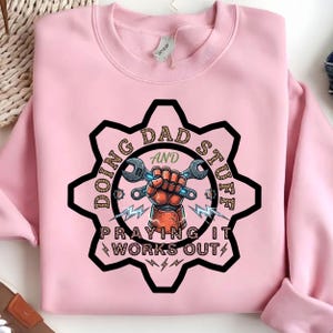 May include: A light pink crewneck sweatshirt featuring a graphic of a gear shape containing a clenched fist holding two wrenches. The text reads "DOING DAD STUFF AND PRAYING IT WORKS OUT" in a leopard print pattern, accented by small lightning bolt icons.
