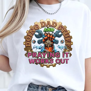 May include: A white t-shirt featuring a graphic of a mechanical fist holding a wrench inside a gear. The text reads DOING DAD STUFF AND PRAYING IT WORKS OUT. The design includes leopard print lettering, blue lightning bolts, and a central star emblem.