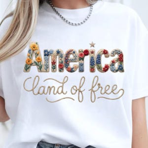 May include: A white t-shirt featuring the word America in large, block letters filled with colorful floral patterns and a small star. Below, the phrase land of free is written in a tan, rope-like cursive script across the chest of the garment.