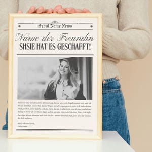 May include: A framed black and white photograph of a woman in a graduation cap. The frame has text at the top that reads "Schul Name News" and "Name der Freundin SHSIE HAT ES GESCHAFFT!" in a newspaper-like layout. The frame is held by a person.