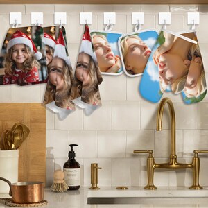 Può includere: Accessori da cucina appesi a ganci bianchi. Include una foto a tema natalizio di un bambino con un cappello da Babbo Natale, due asciugamani con il volto di un bambino, due presine quadrate e un guanto da forno con il volto di una donna. Un rubinetto dorato è in primo piano.