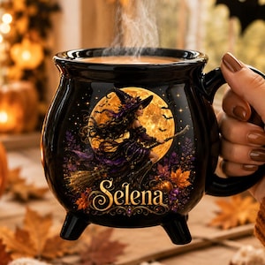 May include: A black ceramic cauldron-shaped mug with three small feet and a handle. The front features a graphic of a witch flying on a broomstick against a large yellow full moon, surrounded by autumn leaves and the name Selena in gold script.