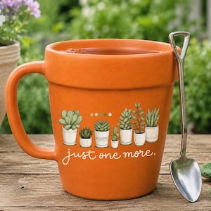Könnte beinhalten: Ein orangefarbener Keramikbecher in Form eines Terrakotta-Blumentopfs mit Henkel. Die Vorderseite zeigt eine Grafik mit sechs Sukkulenten in Töpfen und den weißen Schriftzug "just one more". Ein silberner Metalllöffel in Schaufelform liegt daneben.