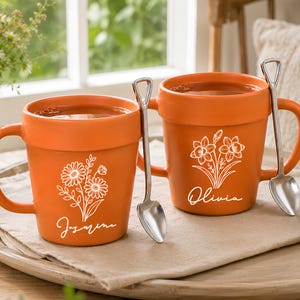 May include: Two terracotta-colored ceramic mugs shaped like flower pots, each featuring a white line-art floral illustration and a personalized name in script. One mug displays the name Jasmine, the other Olivia. Each mug includes a silver shovel-shaped spoon.