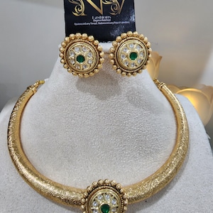 May include: A gold-coloured jewellery set comprising a necklace and earrings. The necklace is a textured, curved band with a central circular pendant. The earrings and pendant feature a central green stone surrounded by smaller white stones and a beaded border. The brand name is "NV Fashions".