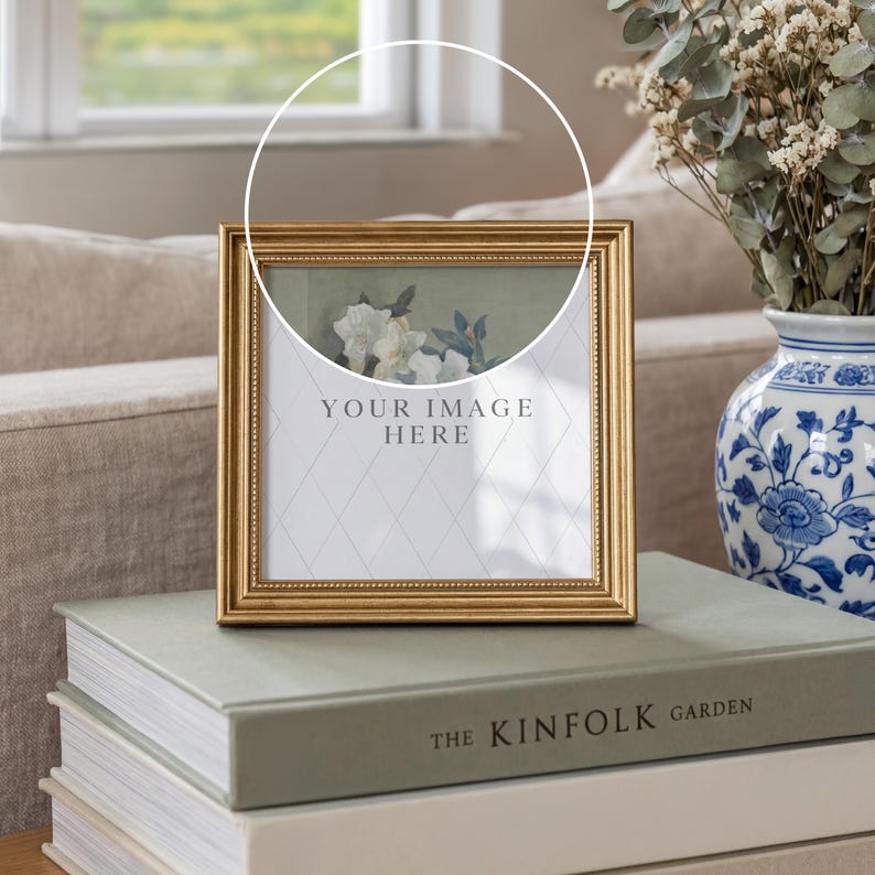 May include: A gold-framed picture frame with the text "YOUR IMAGE HERE" is displayed on a stack of books. The frame has a decorative border and a floral print. A blue and white vase with flowers is in the background.