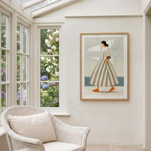 Puede incluir: Una impresi&oacute;n art&iacute;stica enmarcada representa a una persona con un vestido a rayas caminando cerca del mar. La obra de arte presenta una paleta neutra con rayas grises y blancas, zapatos naranjas y un mar azul. La impresi&oacute;n est&aacute; enmarcada en un marco de madera clara.
