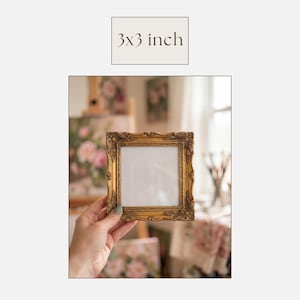 May include: A gold-colored, ornate picture frame, measuring 3x3 inches, is held in a hand. The frame has a square opening and detailed scrollwork. The text "3x3 inch" is above the frame. The background includes a painting and a table.