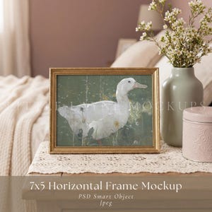 May include: A horizontal 7x5 inch gold picture frame containing an illustration of a white duck in a green field. The frame is positioned next to a light green vase filled with small white flowers and a small pink lidded container on a lace runner.