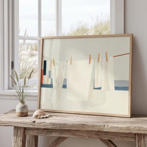 May include: A framed art print showing white linen sheets hanging on a washing line, secured with wooden pegs. The artwork uses a minimalist style with soft colours, including white, blue, and beige. The frame is made of light-coloured wood.
