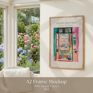 May include: A rectangular A2 frame containing a print of Henri Matisse's Open Window, featuring a colorful painting of an open window overlooking a harbor. The frame is light wood and is mounted on a plain wall next to a window with garden views.