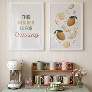 May include: Two framed wall prints, one with the text THIS KITCHEN IS FOR Dancing and one with lemon illustrations. A cream espresso machine, a wooden shelf with eight assorted mugs, two clear glasses, and a small vase of flowers.