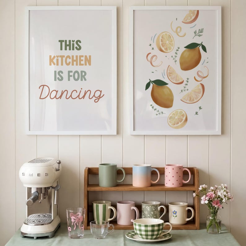 May include: Two framed wall prints, one with the text THIS KITCHEN IS FOR Dancing and one with lemon illustrations. A cream espresso machine, a wooden shelf with eight assorted mugs, two clear glasses, and a small vase of flowers.