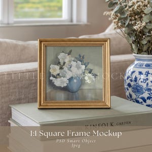 May include: A square gold frame mockup with a painting of white flowers in a blue vase. The frame rests on a stack of books with the text "THE KINFOLK GARDEN" and "1:1 Square Frame Mockup". A blue and white vase with flowers is in the background.