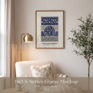 May include: A rectangular wall art print in a thin brown frame featuring a blue and white floral pattern with a central circular motif and the text William Morris Wall Paper Collection, displayed on a light wall above a white armchair.