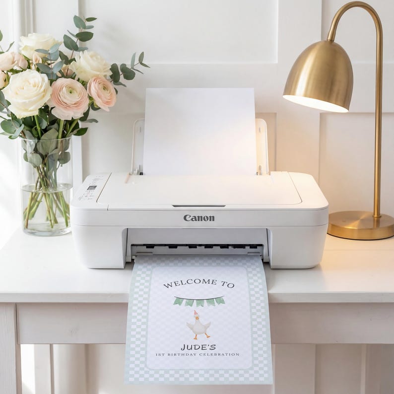 May include: A white Canon printer is printing a welcome sign for Jude's 1st birthday celebration. The sign features a goose illustration and banner. A bouquet of flowers and a gold lamp are also in the image.