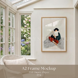 Puede incluir: Maqueta de marco A2 con una ilustración de una persona leyendo un libro, con un suéter verde y pantalones rojos. La obra de arte está en un marco de madera clara, en una pared blanca. La imagen está en una habitación luminosa con una ventana y una silla de mimbre.