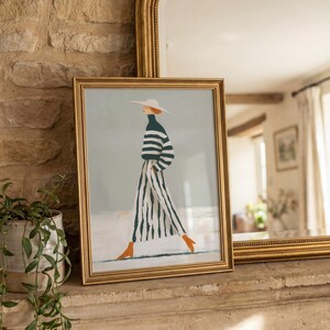 May include: A framed art print featuring a fashion illustration of a person in a striped outfit and hat, walking. The artwork has a gold frame and is displayed on a stone ledge. The person is wearing a green and white striped top and pants, with orange shoes.