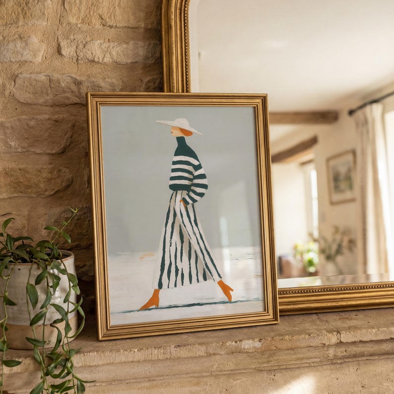 May include: A framed art print featuring a fashion illustration of a person in a striped outfit and hat, walking. The artwork has a gold frame and is displayed on a stone ledge. The person is wearing a green and white striped top and pants, with orange shoes.